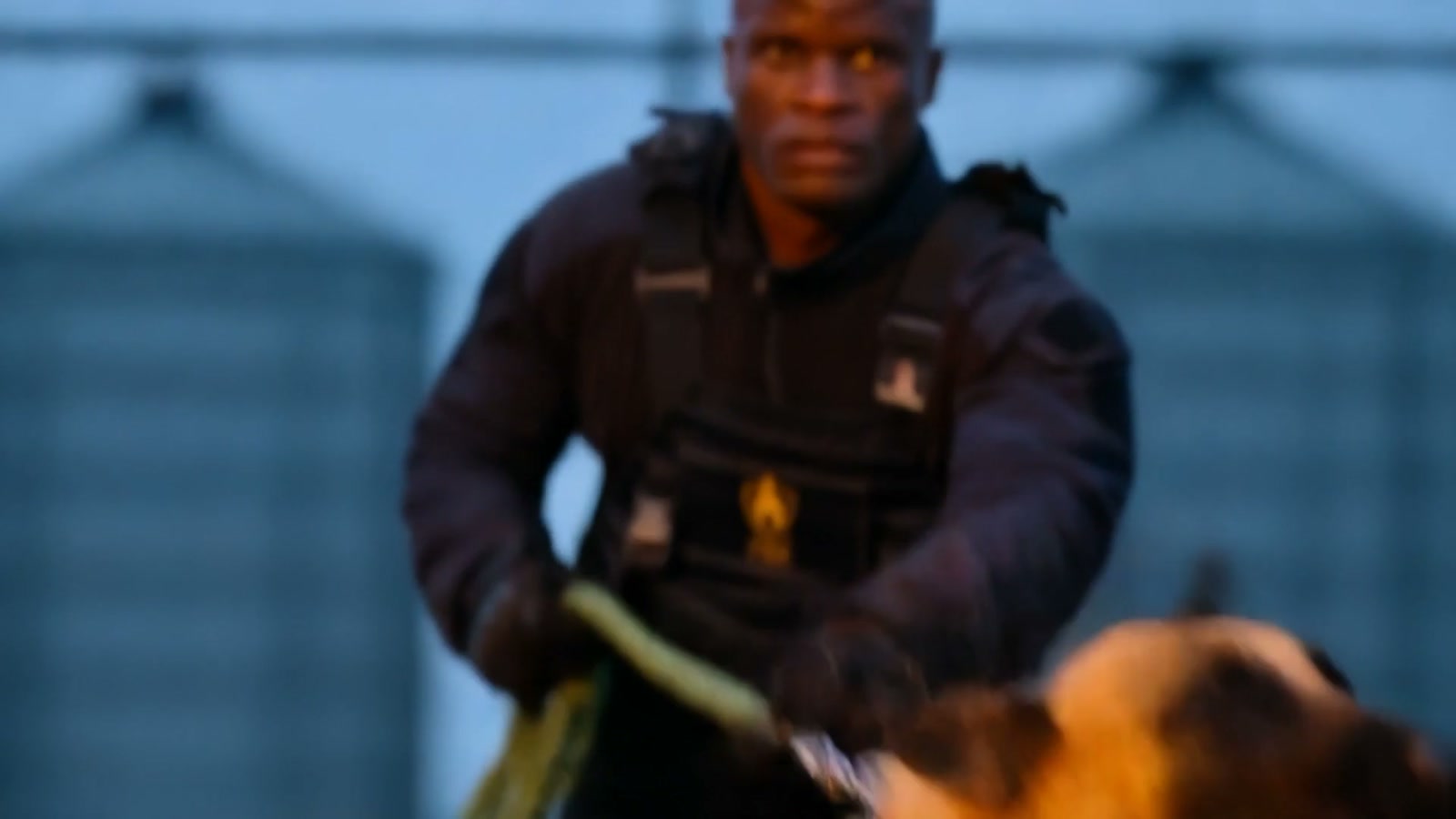 Scene 03 Hunt - K9 handler with Belgian Malinois at razor wire perimeter at blue hour with grain silos